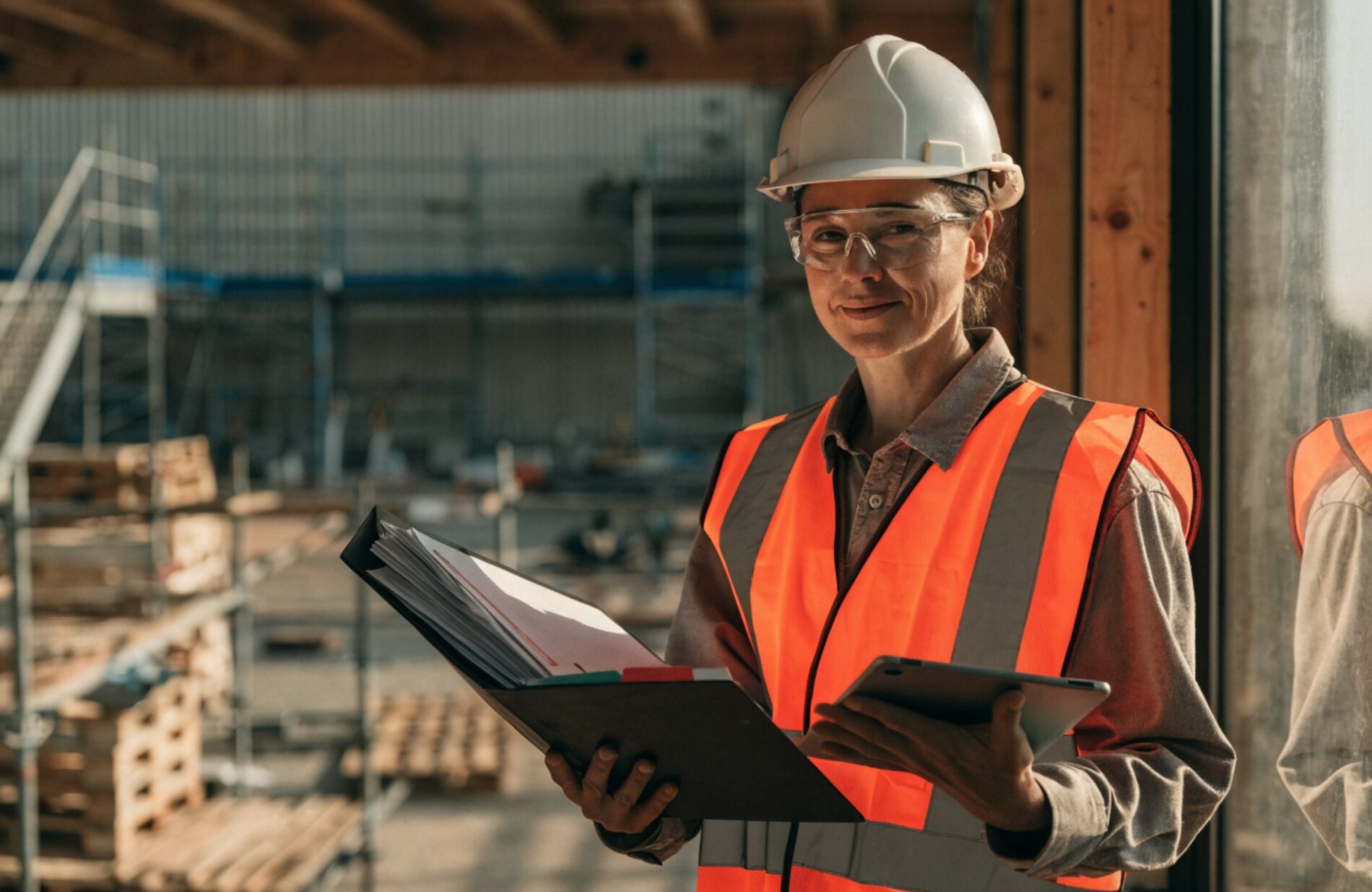 A safety manager in a hard hat, safety glasses, and high-visibility vest reviewing a binder and tablet on a clean job site